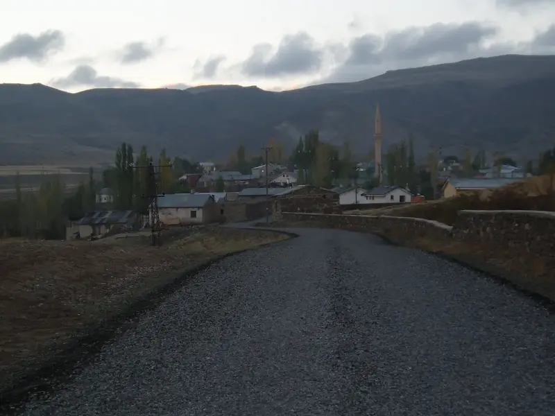 Dağların eteğindeki köye uzanan mıcırlı yol, evler, kavaklar ve cami minaresi